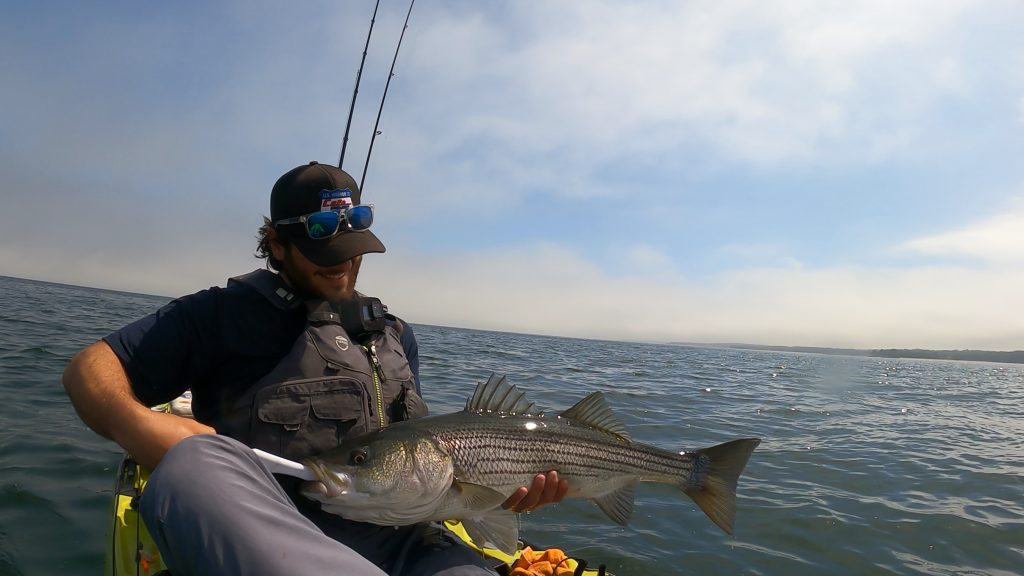 A kayak angler holding a striped bass. The ocean is visible in the background.